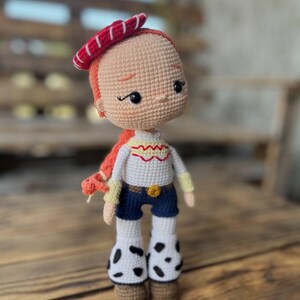 May include: A crocheted doll with red hair, a red and white striped hat, a white shirt with a yellow embroidered design, blue jeans, and white and black cow print boots.