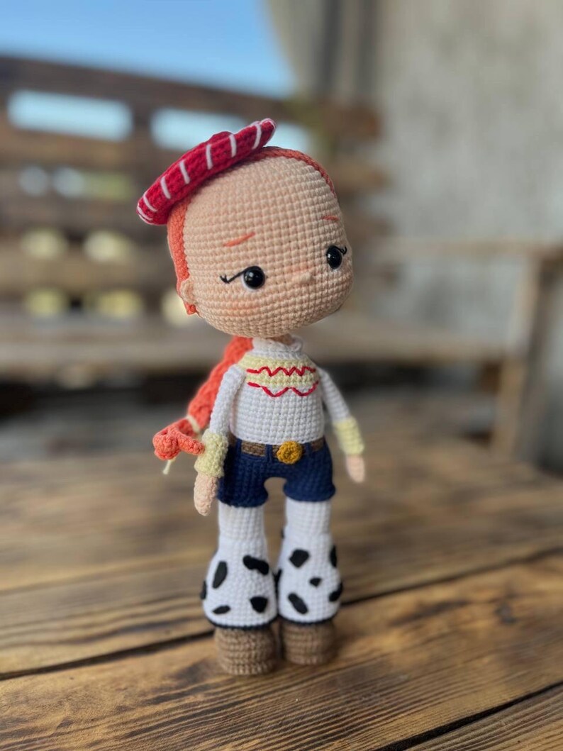 May include: A crocheted doll with red hair, a red and white striped hat, a white shirt with a yellow embroidered design, blue jeans, and white and black cow print boots.