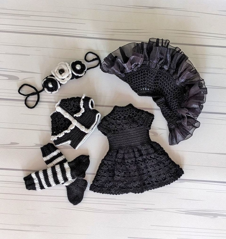 May include: A black crocheted dress, jacket, and leg warmers with white accents. A black and white crocheted headband with white flowers is also included.