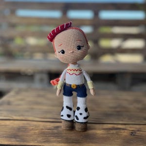 May include: A crocheted doll with red hair, a red and white hat, a white shirt with yellow and red accents, blue jeans, and white boots with black spots. The doll is standing on a wooden surface.