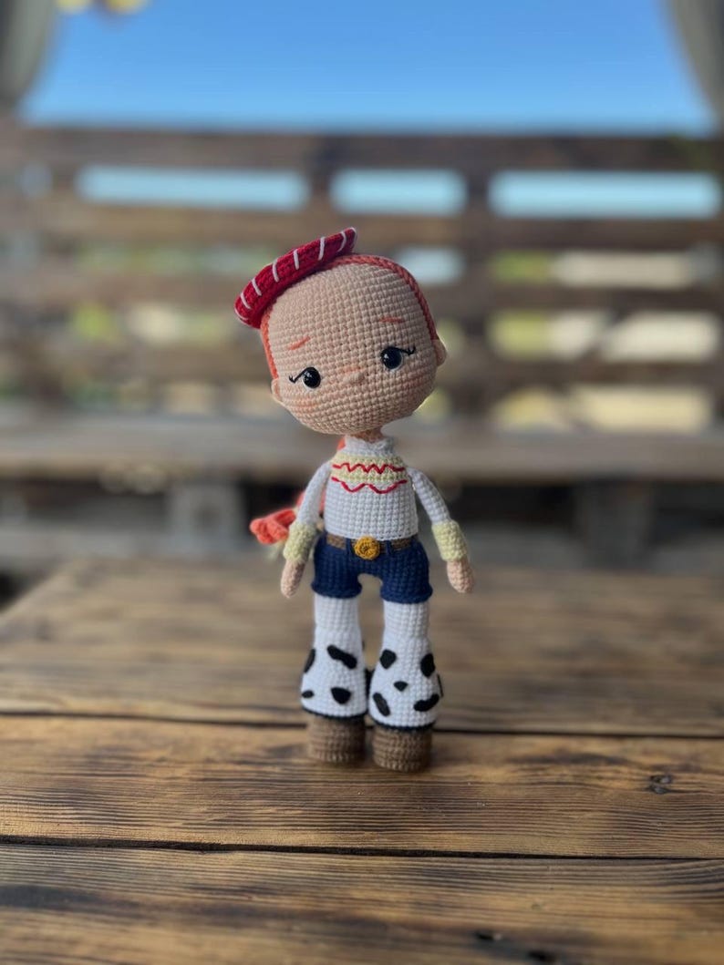 May include: A crocheted doll with red hair, a red and white hat, a white shirt with yellow and red accents, blue jeans, and white boots with black spots. The doll is standing on a wooden surface.