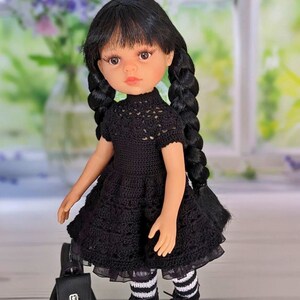 May include: A black crochet dress with a lace trim, worn by a doll with black hair in braids. The doll is wearing black and white striped tights and black shoes. The doll is holding a black backpack.