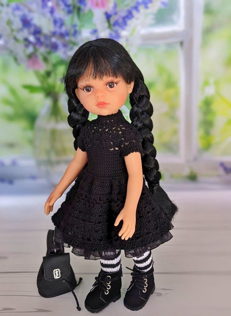 May include: A black crochet dress with a lace trim, worn by a doll with black hair in braids. The doll is wearing black and white striped tights and black shoes. The doll is holding a black backpack.