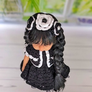 May include: A black and white crocheted doll with long black hair in a braid. The doll is wearing a black dress with white crocheted trim and a white crocheted flower headband with black accents.