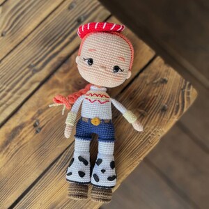 May include: A crocheted doll with red hair, a red and white hat, a white shirt with a yellow design, blue jeans, and brown boots. The doll is wearing a white and black spotted skirt.