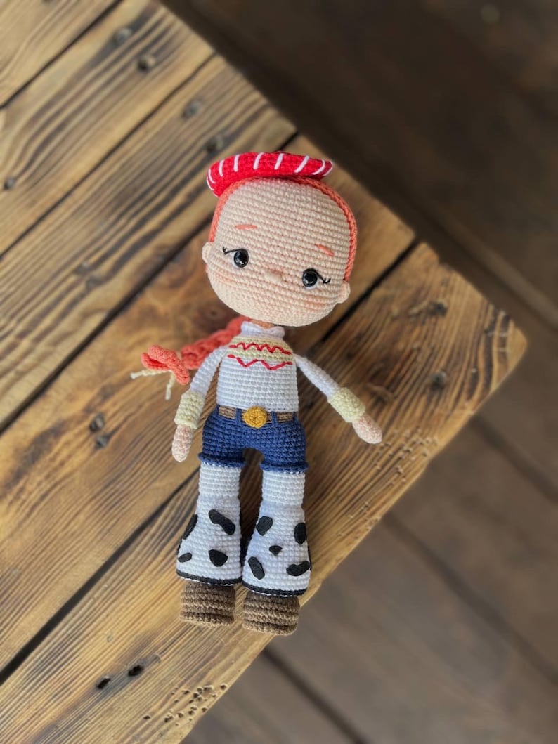 May include: A crocheted doll with red hair, a red and white hat, a white shirt with a yellow design, blue jeans, and brown boots. The doll is wearing a white and black spotted skirt.