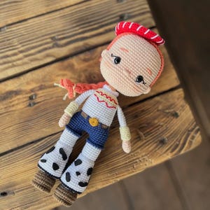 May include: A crocheted doll with red hair, a red and white hat, a white shirt with a yellow star, blue jeans, and white boots with black spots.