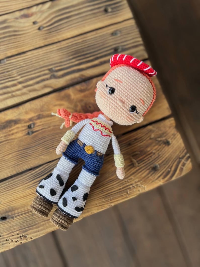 May include: A crocheted doll with red hair, a red and white hat, a white shirt with a yellow star, blue jeans, and white boots with black spots.