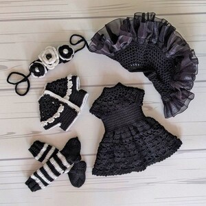 May include: A black crochet baby outfit set featuring a dress, jacket, headband and leg warmers. The headband is adorned with white flowers and pearls. This set is ideal for a baby girl.