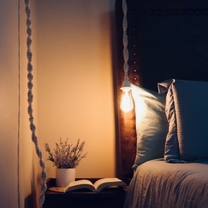 May include: A bedroom scene featuring a hanging light fixture with a visible Edison bulb. A macrame cord suspends the light. A bed with blue pillows and a book on a nightstand are visible. A small potted plant adds a touch of nature.