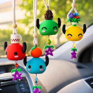 Peut inclure: Quatre ornements de voiture suspendus représentant des personnages arrondis et colorés avec des cornes noires et des visages souriants. Les ornements incluent un rouge avec une tasse, un bleu avec une sphère orange, un vert avec une pomme de pin et un jaune avec des fleurs.