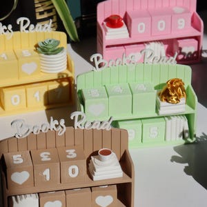 May include: A selection of miniature bookshelves in pastel shades, each labelled "Books Read". The shelves feature numbered blocks and small decorative items. These shelves are designed to track the number of books read, with measurements in cm.