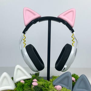 Puede incluir: Auriculares blancos con orejas de gato rosas en un soporte negro. También hay cuatro juegos de orejas de gato adicionales en blanco y gris.