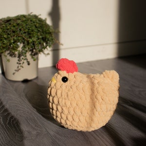 May include: A crocheted plush toy shaped like a chicken. The chicken is beige with a red comb and black eyes. The chicken is sitting on a wooden floor.