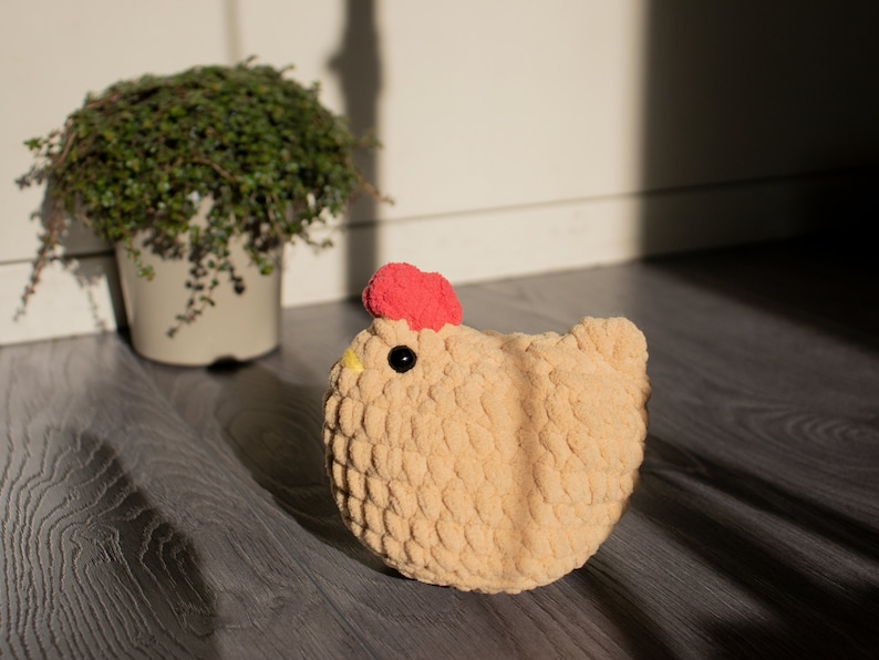 May include: A crocheted plush toy shaped like a chicken. The chicken is beige with a red comb and black eyes. The chicken is sitting on a wooden floor.