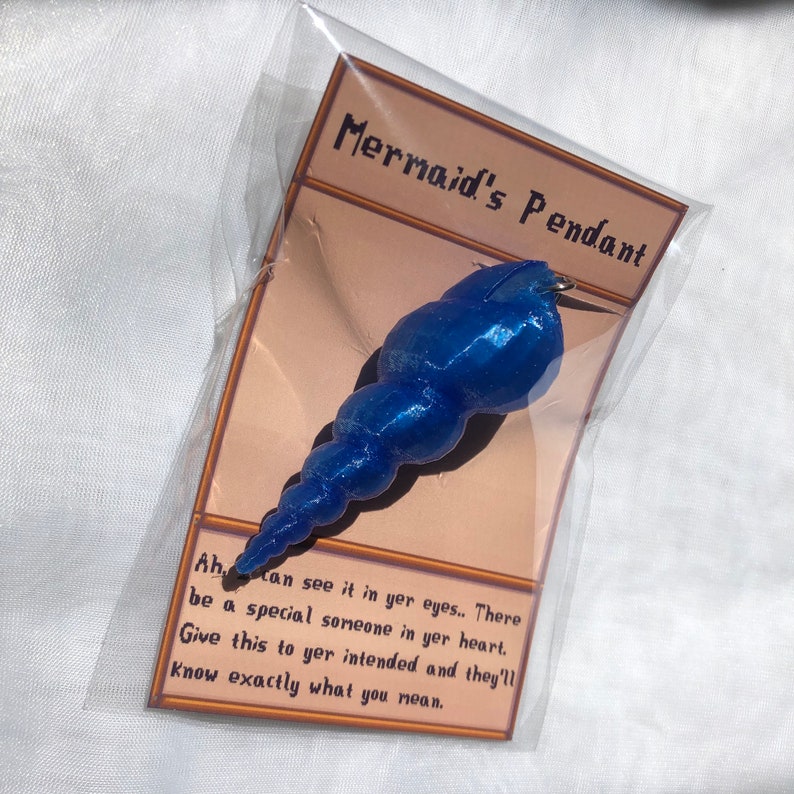Op de afbeelding: Een blauwe zeemeermin-hanger in de vorm van een schelp. De hanger is verpakt in een doorzichtige plastic zak met een kaart waarop "Mermaid's Pendant" en "Ah can see it in yer eyes.. There be a special someone in yer heart. Give this to yer intended and they'll know exactly what you mean." staat.