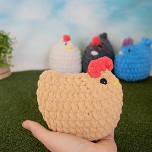 May include: A collection of crocheted chicken plushies. The plushies are in various colors, including white, black, blue, and tan. The tan chicken is being held in a hand.