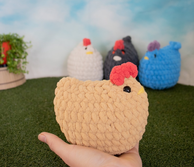 May include: A collection of crocheted chicken plushies. The plushies are in various colors, including white, black, blue, and tan. The tan chicken is being held in a hand.