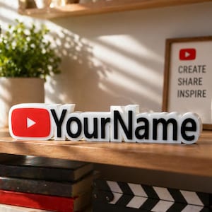 May include: A 3D desk sign featuring the red and white YouTube play button icon followed by the black text YourName in a bold sans-serif font. The sign is constructed from white material with black lettering and a red icon on the left side.