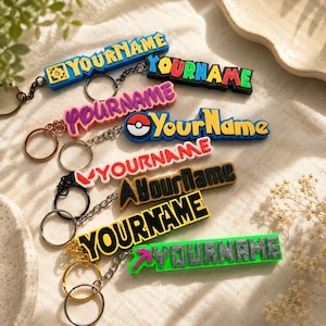 May include: A selection of colourful keychains, each displaying the text "YOURNAME" in diverse fonts and designs. Keychains include blue, black, pink, yellow, red, and green, with attached key rings and chains. The keychains are made of a rubbery material.