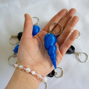 Op de afbeelding: Een blauwe schelp sleutelhanger met een glinsterende afwerking. De sleutelhanger is bevestigd aan een zilveren ring en wordt vastgehouden in een hand die een parel armband draagt.