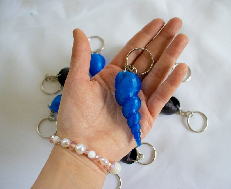 Op de afbeelding: Een blauwe schelp sleutelhanger met een glinsterende afwerking. De sleutelhanger is bevestigd aan een zilveren ring en wordt vastgehouden in een hand die een parel armband draagt.