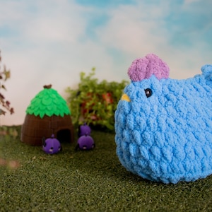 May include: A blue crocheted chicken toy with a purple tuft on its head. The chicken is sitting on a green grassy surface with a small brown house and purple toy bugs in the background.