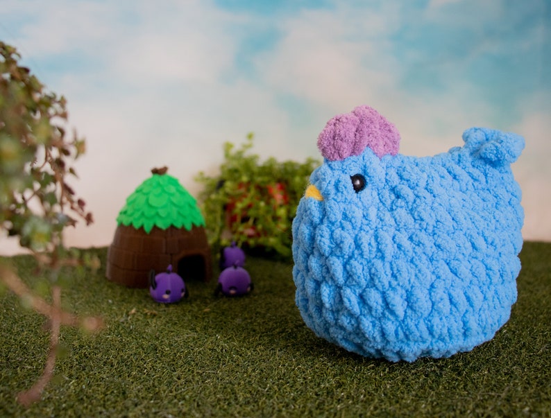 May include: A blue crocheted chicken toy with a purple tuft on its head. The chicken is sitting on a green grassy surface with a small brown house and purple toy bugs in the background.