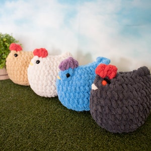 May include: Four crocheted chicken plushies in different colors: brown, white, blue, and gray. The plushies are arranged in a row on a green surface.