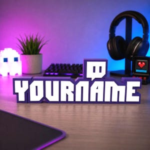 May include: A custom 3D printed sign featuring the purple and white Twitch logo icon followed by the text YOURNAME in a bold, blocky font. The sign is constructed with a deep purple base and raised white lettering, designed for display on a desk.