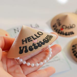 May include: A beige conical hat-shaped charm with the words "HELLO Vietnam" in black, a pink heart, and a white beaded chain. Other charms with Vietnamese text are visible.
