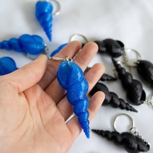 Op de afbeelding: Een close-up van een hand die een blauwe schelp sleutelhanger vasthoudt. De schelp is gemaakt van plastic en heeft een glanzende, iriserende afwerking. Er zijn andere schelp sleutelhangers op de achtergrond, sommige zijn blauw en sommige zijn zwart.