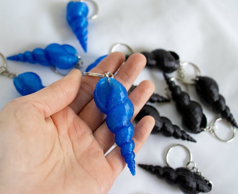 Op de afbeelding: Een close-up van een hand die een blauwe schelp sleutelhanger vasthoudt. De schelp is gemaakt van plastic en heeft een glanzende, iriserende afwerking. Er zijn andere schelp sleutelhangers op de achtergrond, sommige zijn blauw en sommige zijn zwart.