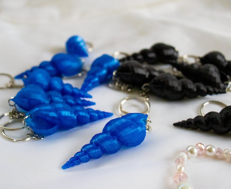 Op de afbeelding: Een set blauwe schelp sleutelhangers met zilveren ringen. De schelpen zijn in een rij gerangschikt, met de grootste schelp aan het einde van de rij. De schelpen zijn allemaal van verschillende grootte en vorm.