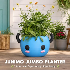 May include: A bright blue, round planter shaped like a Junimo character with two black side handles, two large black circular eyes, and two small pink oval cheeks. The planter is filled with green leafy plants and yellow flowers.