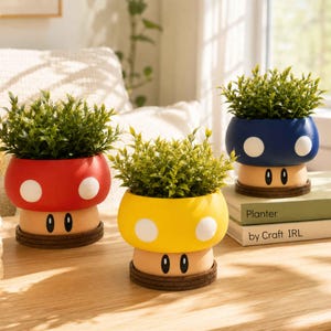 Puede incluir: Tres macetas con forma de hongo en rojo, amarillo y azul, cada una con lunares blancos y una cara de dibujos animados. Las macetas contienen plantas verdes de hojas y están sobre una superficie de madera. Las macetas miden unos 13 cm de alto.
