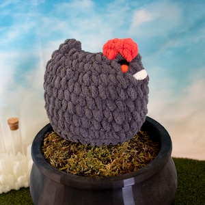 May include: A gray crocheted chicken toy with a red comb and a white beak sits in a black pot filled with green moss.