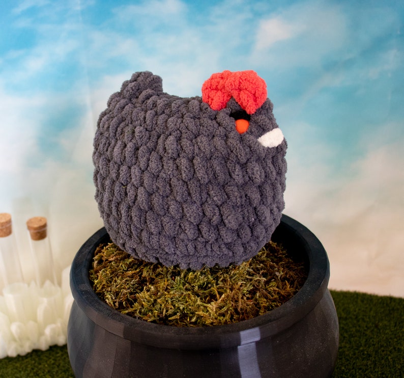 May include: A gray crocheted chicken toy with a red comb and a white beak sits in a black pot filled with green moss.