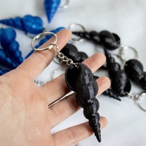 Op de afbeelding: Een zwarte schelp sleutelhanger met een zilveren ketting en een zilveren ring. De sleutelhanger is gemaakt van een glad, glanzend materiaal en heeft een spiraalvorm.