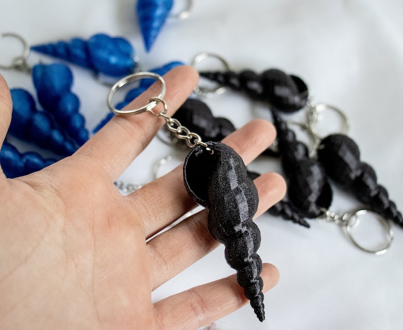 Op de afbeelding: Een zwarte schelp sleutelhanger met een zilveren ketting en een zilveren ring. De sleutelhanger is gemaakt van een glad, glanzend materiaal en heeft een spiraalvorm.