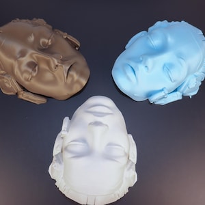 May include: Three sculpted face busts in bronze, light blue, and white. The busts are arranged on a dark surface. Each face has closed eyes and detailed features, creating a classical aesthetic.