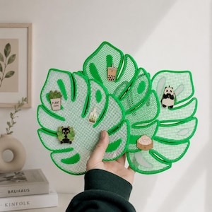 May include: A green, leaf-shaped memo board with a white mesh design. The board is decorated with several small, decorative pins, including a black cat, a panda, and a plant. The text "Plant Lover" is visible on one of the pins.