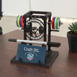 May include: Black barbell-shaped business card holder with colourful weight plates and the text "PERSONAL TRAINER". Includes a silver pen and a business card with the text "Craft IRL". A small succulent plant in a black pot is also present.