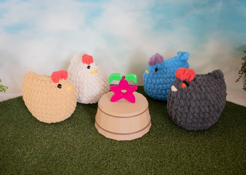 May include: Five crocheted plush chickens in different colors, including beige, white, blue, and gray, are arranged on a green surface. A pink star-shaped object is in the center of the image, sitting on a wooden base.