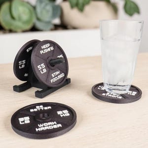 May include: Black weight plate coasters with motivational phrases and weight measurements in kilograms. A clear glass of water sits on one coaster. A weight plate holder with two plates is also present.