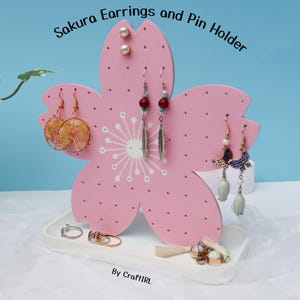 以下が含まれることがあります： 「Sakura Earrings and Pin Holder」の文字が入った、ピンク色の花の形をしたイヤリングとピンのホルダー。ビーズや蝶のデザインなど、さまざまなイヤリングを展示しています。ベースの白いトレイには、指輪やその他の小さなジュエリーが置かれています。