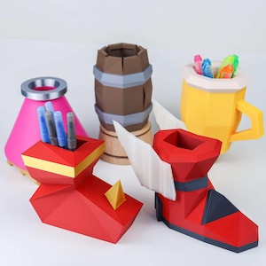May include: A collection of colorful, geometric-shaped desk organizers. Includes a pink conical container, a red and yellow pen holder, a brown barrel-shaped container, a yellow mug, and a red boot-shaped container with white wings. Each container holds pens or markers.