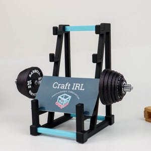 May include: A black and light blue barbell-themed business card holder with the text "Craft IRL" on a card. The holder has a weight on each end, with one weight labeled "25 KG".