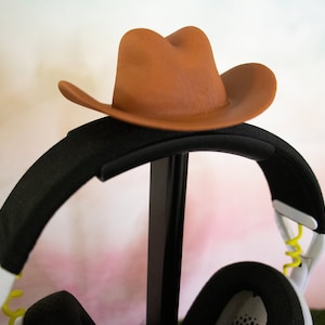 Può includere: Un cappello da cowboy marrone si trova sopra un supporto per cuffie nero. Le cuffie sono bianche con accenti gialli e un cavo a spirale.