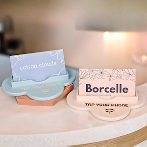 Op de afbeelding: Twee wolk-vormige visitekaarthouders. Eén is lichtblauw met een roze basis en houdt een kaart met "cotton clouds". De andere is gebroken wit en houdt een kaart met "Borcelle". Beide houders hebben "TAP YOUR PHONE" erop gedrukt.